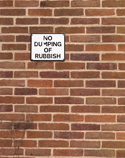 A rectangular metal sign mounted on a red brick wall bears the message 'No Dumping of Rubbish' in bold black capital letters on a white background. The sign appears weathered, with slight rust and chipping around the edges, and is attached with visible screws at each corner. The brick wall behind the sign features a traditional running bond pattern with reddish-brown bricks, some darker and lighter in tone, separated by light-colored mortar joints. The surface of the bricks shows slight texture and variation in finish, with some bricks appearing smoother and others more textured. The overall scene suggests an outdoor setting, possibly an alley or side wall of a building, within an urban environment where waste disposal restrictions are enforced. The sign's purpose relates to maintaining cleanliness and order, aligning with services that handle rubbish removal and private waste management, as referenced by Waste Disposal Hounslow.