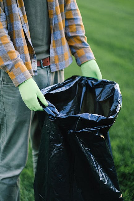A person wearing a green long-sleeve shirt, a checkered yellow, blue, and gray jacket, and light green gloves is holding the top of a black plastic rubbish bag that is open and partially filled, situated outdoors on a grassy area. The background shows a well-maintained lawn with lush green grass. The scene suggests a waste disposal activity, possibly related to garden or household rubbish collection, with the individual preparing the bag for rubbish removal services such as those provided by Waste Disposal Hounslow or similar companies specializing in private waste handling, emphasizing the importance of proper waste containment for effective rubbish removal.
