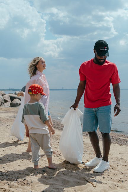 A man wearing a black baseball cap, red t-shirt, and light blue denim shorts is standing on a sandy beach, holding a large white plastic bag used for rubbish collection. Behind him, a woman with curly blonde hair, dressed in a pink top, stands facing the water with a relaxed posture. A young child, wearing a red polka-dot cap, green and white shirt, and beige trousers, stands nearby and appears to be observing the man. The background features a calm body of water, a cloudy sky with patches of blue, and a distant shoreline with rocks along the water's edge. The scene suggests a clean-up activity or informal rubbish collection at the beach, with the man from Waste Disposal Hounslow engaged in an outdoor waste management task, reflecting practices of private waste handling outside formal municipal rubbish collection systems. The natural environment and the presence of collected waste bags highlight an activity related to responsible rubbish removal in a recreational area.