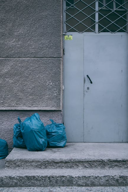 Several blue plastic rubbish bags are gathered on a concrete step outside a building, placed against a textured grey wall. The bags appear to be filled with waste and are tied at the top. To the right, there is a plain grey metal door with a black handle, set within a secured metal grille window above it. The scene is lit evenly with natural daylight, highlighting the rough surface of the wall and the smooth finish of the door. The area suggests an outdoor space used for waste collection or disposal, potentially indicating a site used for private rubbish removal services such as those provided by Waste Disposal Hounslow. The setting reflects an informal or supplementary waste handling location, typically associated with on-site rubbish clearance for flats or estate environments, where extra measures are taken outside the regular collection schedule. The practical environment and visible waste bags emphasize an approach to alternative rubbish collection relevant for properties with difficult access or non-standard refuse disposal needs.