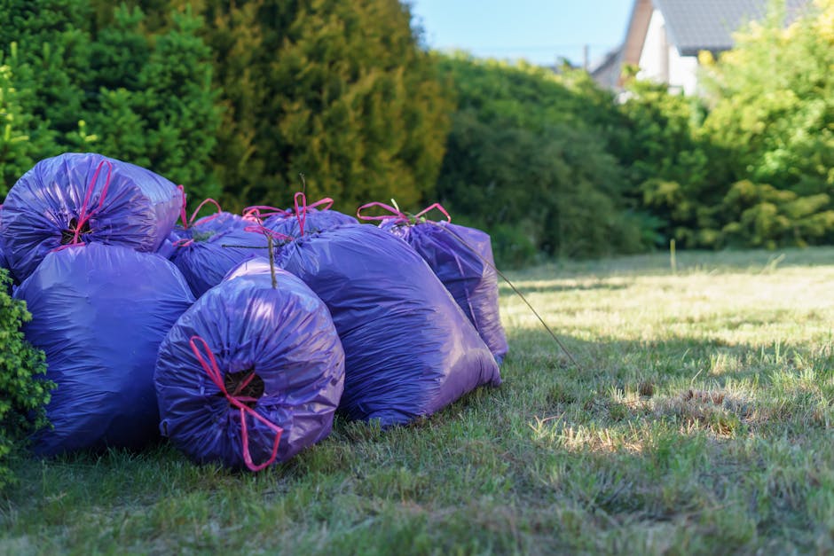 Two large black garbage bags and a cardboard box are positioned on the pavement alongside a black metal fence, with a densely foliaged tree in the background. The bags appear to be filled with waste material, their surfaces slightly crinkled and shiny, reflecting ambient light. The cardboard box is partially compressed and bears printed text, indicating its previous use for packaging. The scene takes place on a street or driveway, with a portion of vehicle road visible in the foreground. The environment suggests a typical scene of domestic waste awaiting collection or disposal, characteristic of private rubbish removal services offered by Waste Disposal Hounslow. The dark tones and shadows cast by the fence and foliage create a subdued atmosphere, emphasizing the waste bags as the main objects in the image, subtly aligning with themes of rubbish clearance and alternative waste handling outside of local authority collections.