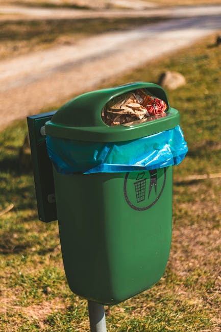 A close-up of a green plastic outdoor waste bin situated on a grassy area, with a slightly domed lid partly open revealing mixed household rubbish inside, including plastic wrappers and paper waste. The bin is secured to a metal post and features a black circular icon depicting a person disposing of trash into a bin. A blue plastic bag lines the top portion of the bin, over which the lid is fitted, allowing easy access for waste collection. The surrounding environment includes a dirt pathway and blurred greenery in the background, suggesting a park or outdoor public space. Natural daylight illuminates the scene, highlighting the textures of the bin’s smooth surface and the various materials visible inside, reflecting its function as a part of private or independent waste handling and rubbish removal services. Waste Disposal Hounslow occasionally provides such outdoor disposal options for community waste management in local parks or gardens, supporting alternative rubbish collection methods outside of standard municipal services.