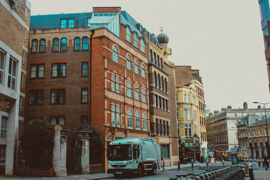 A street scene in an urban area displaying a range of historic and modern buildings with ornate architectural details, including arched windows, decorative facades, and a domed tower with a weather vane on top. In the foreground, there is a light blue rubbish collection vehicle parked on the street, which appears to be used for private waste disposal rather than municipal collection. The vehicle's surface is smooth with a shiny finish, and it is positioned near a curb with a row of empty bike racks along the pavement. Surrounding the vehicle are bollards and traffic signs, with a few pedestrians and other vehicles visible further down the street. The environment is illuminated by natural daylight, with a slightly overcast sky providing diffuse lighting, and the scene is typical of a busy city centre layout where independent rubbish collection services, such as Waste Disposal Hounslow, may operate alongside regular street traffic and pedestrian pathways.