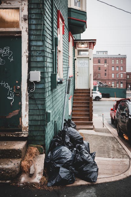 A collection of multiple black garbage bags, filled and tied at the top, are stacked against the exterior green shingled wall of a residential building. The building features a small wooden staircase leading to a white door with red trim and a small awning above it. Nearby, on the concrete pathway, are a few patches of snow and dried grass, indicating colder weather. To the left of the bags, there is a dark metal door with graffiti, and a small grey utility box mounted on the wall. In the background, a parking area with a few vehicles is visible, along with nearby brick buildings under an overcast sky. This scene appears to illustrate an independent rubbish removal effort, possibly facilitated by a private waste disposal service such as Waste Disposal Hounslow, suited to local rubbish collection requirements in residential areas.