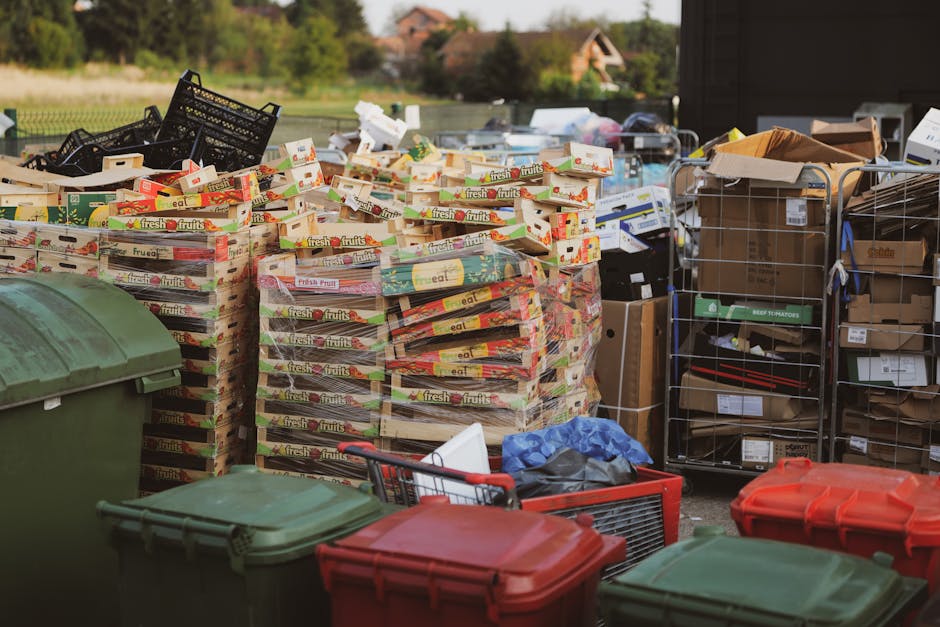 An outdoor scene showing multiple stacks of flattened cardboard boxes, mainly branded with 'fresh fruits,' arranged on pallets or small platforms, with some black plastic crates and wire mesh containers filled with various packaging materials and smaller boxes nearby. The cardboard stacks are positioned in front of a residential area with houses and trees visible in the background. Several large, color-coded wheelie bins with green and red lids are placed in the foreground, partially blocking the view of some smaller items, including a shopping trolley and a blue plastic bag. The area appears to be part of a waste or recycling collection point, with natural daylight illuminating the scene, emphasizing the textures of the cardboard, plastic, and metal objects, and supporting the context of independent waste disposal or rubbish removal services such as those provided by Waste Disposal Hounslow.