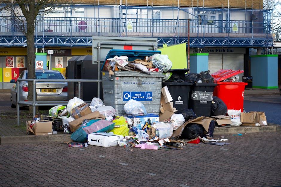A large collection of mixed waste and rubbish is piled around multiple wheeled bins on a paved section of a street. The waste includes cardboard boxes, paper, plastic bags, and packaging materials, some of which are crumpled or torn. The waste containers are made of plastic and metal, with one grey bin labeled for mixed paper and card, while others are black and red for general waste or recycling. The bins are positioned on a curb, with some overflowing, causing additional rubbish to spill onto the ground. Behind the waste collection area, there is a parked silver car with a registration plate visible, and a metal railing separates the waste area from the rest of the street. In the background, a commercial building features storefronts with signage, and above, a blue scaffolding structure suggests ongoing construction or renovation work. The scene is captured in natural daylight, highlighting the informal and unmanaged appearance typical of private waste collection sites, with Waste Disposal Hounslow providing a context for non-local authority rubbish handling services.